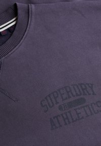 Lilla sweatshirt lavet af blødt materiale, med en ribbet rund halsudskæring og et trykt "Superdry Athletics" logo på forsiden.