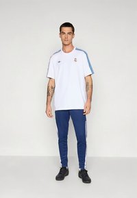 Balti trumpomis rankovėmis marškinėliai mėlynomis juostelėmis ir logotipu, derinami su mėlynomis siaurėjan�čiomis kelnėmis, kurių šonuose baltos juostelės, ir juodais sportbačiais.