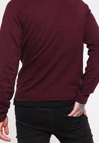 Burgundy stickad tröja med långa ärmar och rundad halslinning. Har en texturerad yta och en enfärgad svart fåll längst ner.