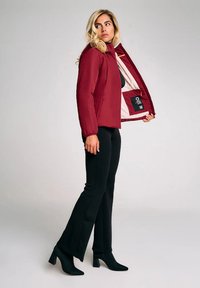 Donna che indossa pantaloni neri e stivali mentre apre una giacca rossa per mostrare la sua leggera fodera interna e la tasca interna con etichetta.