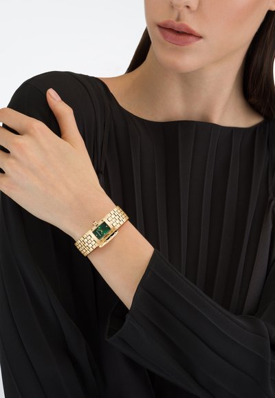 Femeie purtând un top plisat negru, care arată un ceas aurit cu cadran pătrat verde pe încheietura mâinii stângi, pe un fundal alb.