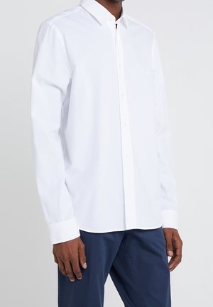 Mężczyzna ubrany w białą koszulę z długim rękawem na guziki i ciemnoniebieskie spodnie na jednolitym jasnym tle.