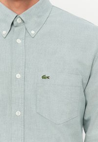 Lys grønn knapp-up skjorte laget av teksturert stoff, med krage, frontlomme og en liten brodert logo.