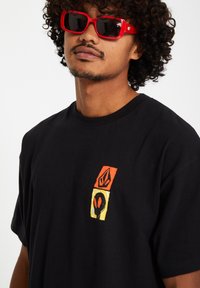 Black cotton t-shirt featuring a small, colorful logo design in orange, yellow, and black on the upper left side.