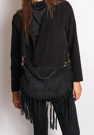 Borsa a tracolla in suede nero con frange, patta arrotondata, intagli decorativi e tracolla regolabile, indossata su un outfit scuro.