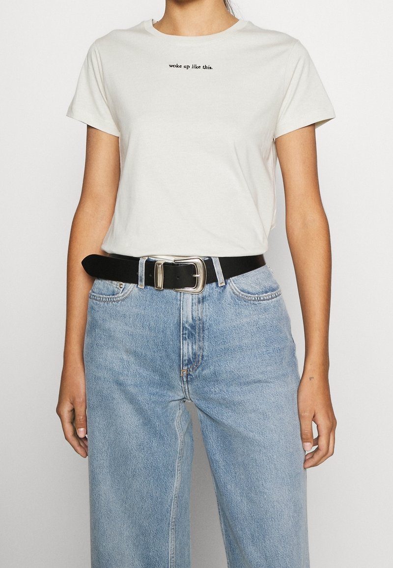 T-shirt en coton couleur crème avec texte noir « woke up like this » ; assorti à un jean bleu clair taille haute et une ceinture noire à boucle argentée.