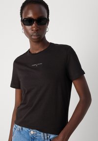 Svart bomull t-skjorte med korte ermer og rund hals, som har en hvit "TOMMY JEANS" logo på brystet. Kombinert med blå denimjeans.