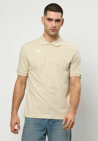 Egy férfi bézs rövid ujjú pólóingben, amelyen logó található a mellrészen és az ujján, kék farmernadrággal, egy egyszínű háttér előtt áll.