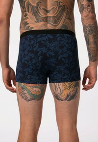 Boxers de color azul marino con un patrón floral, con una cinturilla negra y una textura de algodón suave. Tatuajes visibles en las piernas.