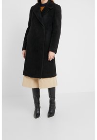 Abrigo largo negro con superficie texturizada, solapas con muesca, cierre de botones y mangas largas, combinado con una falda beige y botas negras hasta la rodilla.