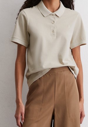 Beige polo skjorte med ribbet krave, tre knapper og et lille broderet logo. Kombineret med brune bukser med vidde i benene.