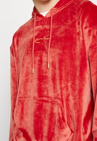 Sudadera de terciopelo rojo con un bolsillo frontal, cordones y un logo bordado en dorado. Textura suave con un corte holgado y capucha.