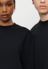 Δύο άτομα φορούν μαύρα μπλουζάκια με στρογγυλή λαιμόκοψη, κομμένα στο επίπεδο των ώμων και των προσώπων, εμφανίζοντας σκουλαρίκια και ουδέτερες εκφράσεις μπροστά σε λευκό φόντο.