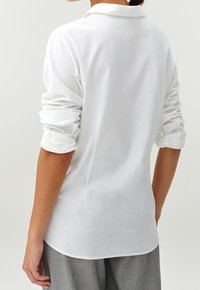 Chemise blanche à manches longues avec une coupe ajustée, texture douce, poignets retroussés et col classique. La vue arrière montre un ourlet droit et des coutures minimales.