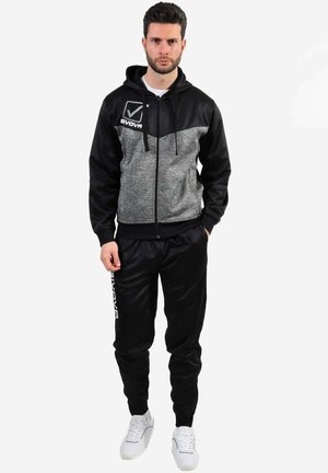 Completo sportivo in nero e grigio con felpa con zip, caratterizzato da un pannello decorato sul petto e pantaloni jogger neri. A completare il look, delle sneaker bianche.
