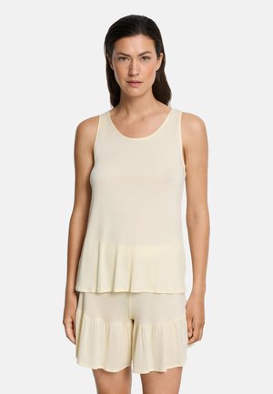 Vrouw die een mouwloze crème top en bijpassende gerimpelde korte broek draagt, staand tegen een effen lichte achtergrond.