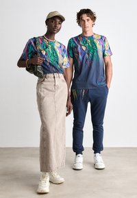 Zwei Personen tragen farbenfrohe Graphic-T-Shirts mit auffälligen Mustern in Grün, Gelb und Blau. Die Frau kombiniert ihr Shirt mit einem beigen Rock, während der Mann marineblaue Jogginghosen trägt. Beide tragen lässiges Schuhwerk.