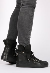 Zapatillas altas negras que combinan cuero texturizado y gamuza, con suelas gruesas, cordones redondos y detalles metálicos.