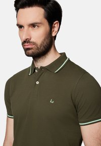 Oliwkowa koszulka polo z teksturowanym wykończeniem, kontrastującymi akcentami w miętowym kolorze przy kołnierzyku i rękawach oraz małym haftowanym logo na piersi.