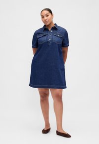 Kortärmad denimklänning i mörk tvätt med knäppning fram, bröstfickor och en avslappnad passform, med en fåll precis ovanför knäna.