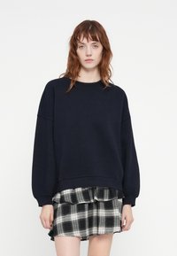 Sweatshirt bleu marine avec des manches-ballons, col rond et ourlet côtelé, associé à une jupe à volants en tartan noir et blanc. Texture lisse.