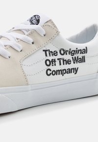 Sneaker med vit canvasöverdel och beiga mockadetaljer. Har svart text som säger "The Original Off The Wall Company" och vita snören.