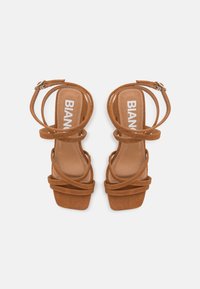Sandales à talons hauts en daim marron avec plusieurs brides entrecroisées, bout carré et semelle intérieure texturée ; le nom de la marque "BIANCA" est inscrit sur la semelle.