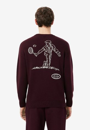 Homme portant un pull bordeaux foncé avec un contour blanc d’un joueur de tennis frappant une balle dans le dos et un petit logo Lacoste Heritage en dessous.