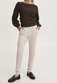 Brown knitted sweater with a chevron pattern, paired with cream striped trousers and black loafers, featuring a slim fit and elastic cuffs.