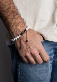 Pulsera de cuentas blancas y negras con un cordón negro texturizado, ajustable con dos cuentas negras, llevada en una muñeca con una camisa de color claro.
