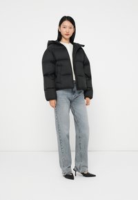 Svart puffjacka med huva, quiltad textur och dragkedjestängning, tillsammans med ljusblå vida jeans och svarta spetsiga klackar.