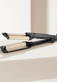 Crni uvijač za kosu s zlatnim cijevima, s postavkama temperature i ergonomskim drškama, odložen na svijetloj teksturiranoj površini.
