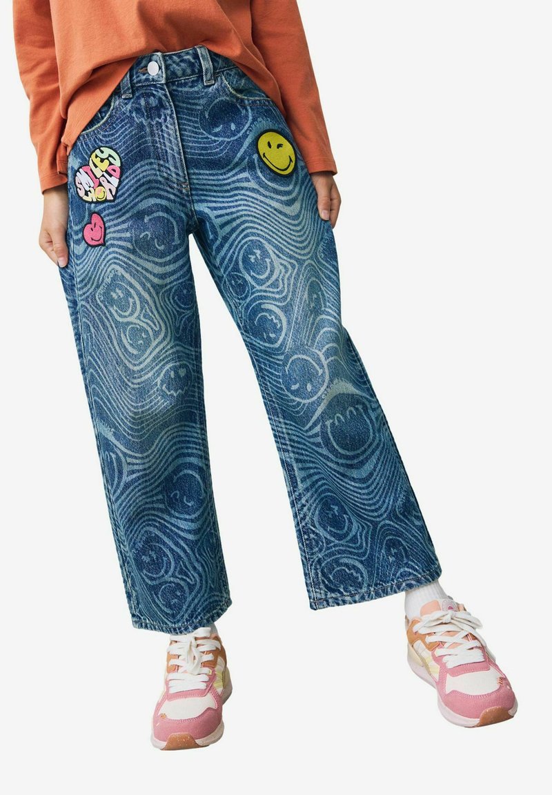 Jean avec motifs ondulés bleus et patchs visage souriant, porté avec des baskets roses et blanches et un t-shirt orange à manches longues.