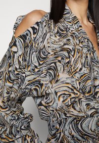 Femme portant un haut à manches longues avec un motif tourbillonnant abstrait noir, blanc et orange et une découpe à l'épaule dénudée d'un côté.