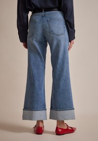 Široke modre jeans hlače iz denima z lahnim denimsko zavihkom, ki imajo dve zadnji žep, skupaj z rdečimi balerinkami. Tekstura se zdi mehka in sproščena.