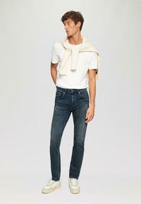 Jeans blu scuro, maglietta bianca semplice, maglione crema poggiato sulle spalle e scarpe da ginnastica chiare. Taglio slim con dettagli minimi.