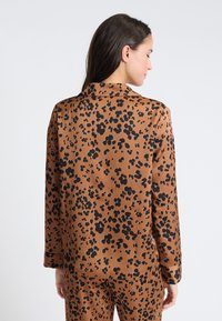 Brązowa bluzka w wzór z czarnymi plamkami lamparta, z długimi rękawami i kołnierzykiem. Gładka tekstura, luźny krój z widocznymi detalami szwów.