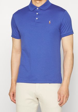 Mand iført en tætsiddende blå kortærmet poloshirt med et lille broderet logo på venstre bryst og beige bukser.