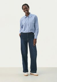 Camisa de botones a rayas azul y blanca con dos bolsillos en el pecho, combinada con unos vaqueros de mezclilla azules sueltos y zapatillas blancas. Diseño sencillo, ajuste casual.
