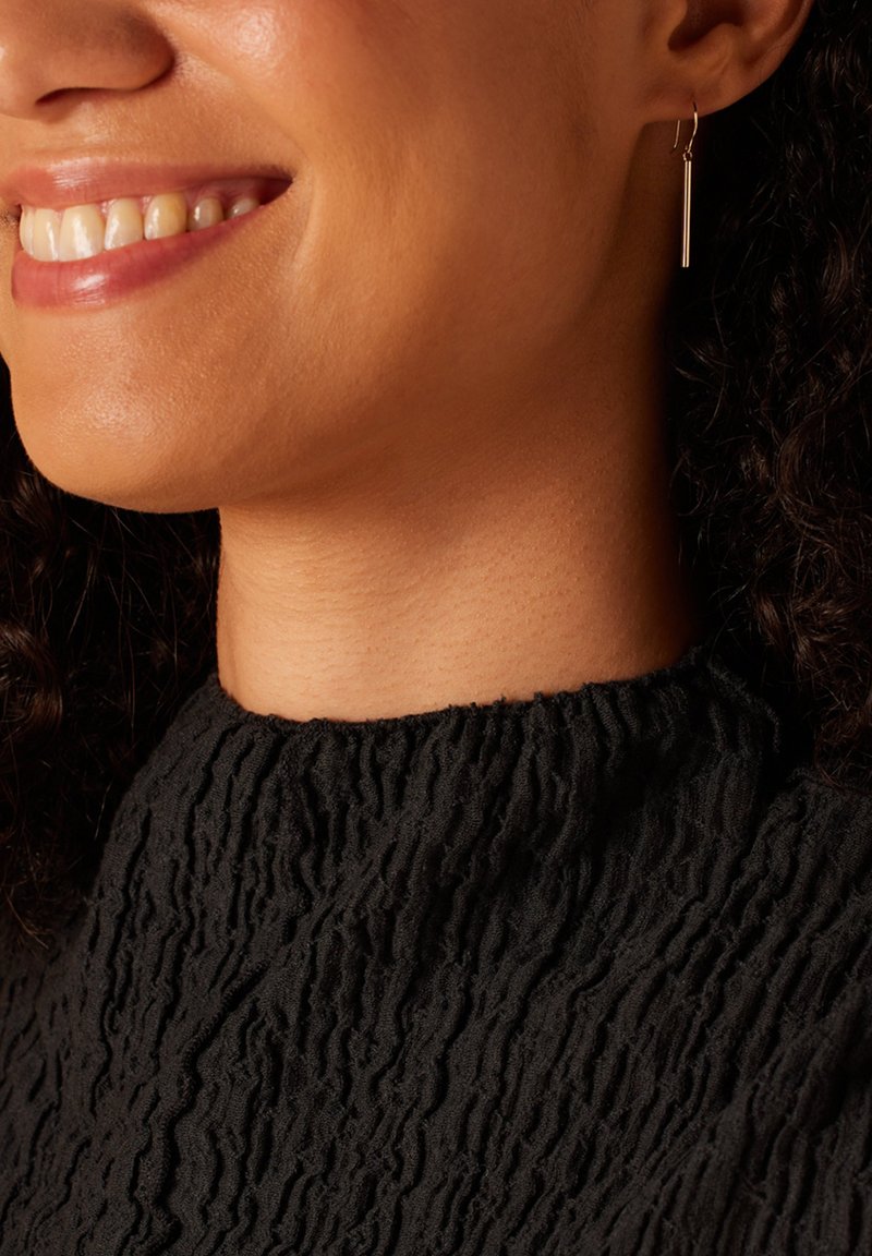 Boucle d'oreille en ton doré avec un détail en barre verticale. En arrière-plan, un vêtement noir texturé avec un col haut et un motif froncé.