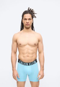 Άνδρας χωρίς μπλούζα με μακριά dreadlocks που φοράει ανοιχτό μπλε εσώρουχα Calvin Klein, στέκεται μπροστά σε απλό λευκό φόντο.