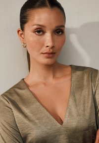 Garām piedurknēm, ar V veida kakla izgriezumu topa, izgatavots no metāliskas olīvzaļas auduma ar gludu tekstūru. Modelis valkā nelielas zelta apaļas auskariņus. Uz ādas redzamas smalkas vasaras raibumi.