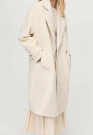 Kvinde iført en lang, off-white uldfrakke med store revers, knapmanchetter og hænder i sidelommerne over en lys kjole.