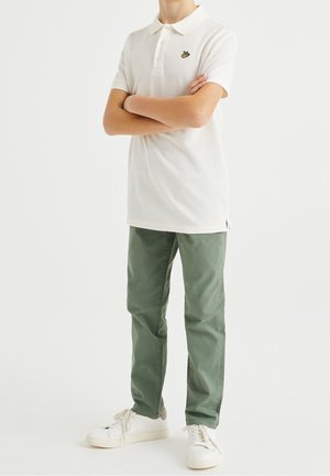 Camisa polo blanca con un logo amarillo, pantalones verdes y zapatillas blancas. El conjunto presenta un corte clásico y una textura suave.