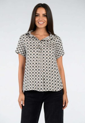 Donna sorridente con lunghi capelli scuri che indossa una camicia a maniche corte, fantasia, con bottoni e pantaloni neri, in piedi davanti a uno sfondo semplice.