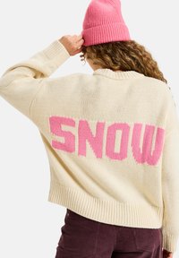 Gebreid sweater in crème met grote roze "SNOW" tekst op de achterkant. Heeft een ronde hals en ribbelmanchetten. Draagt een roze beanie.