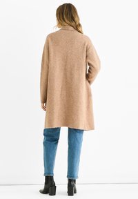 Beige wool-blend long cardigan with a collar, side pockets, and a relaxed fit, paired with light blue jeans and black ankle boots.