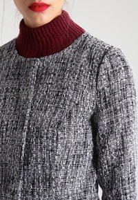 Cappotto texturizzato bianco e nero con un alto colletto a costine bordeaux, caratterizzato da un collo rotondo e chiusura frontale con bottoni.