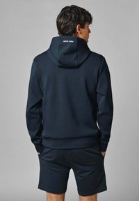 Sudadera marinera con un bolsillo tipo canguro, textura suave, puños acanalados y una gran capucha con un logo en la parte posterior. Combinada con pantalones cortos a juego.