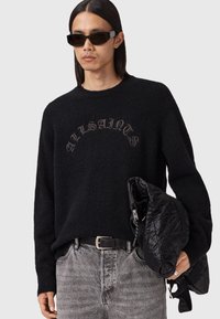 Man wearing black sunglasses and a black sweater with "ALLSAINTS" text, holding a black quilted jacket, dressed in gray washed jeans.